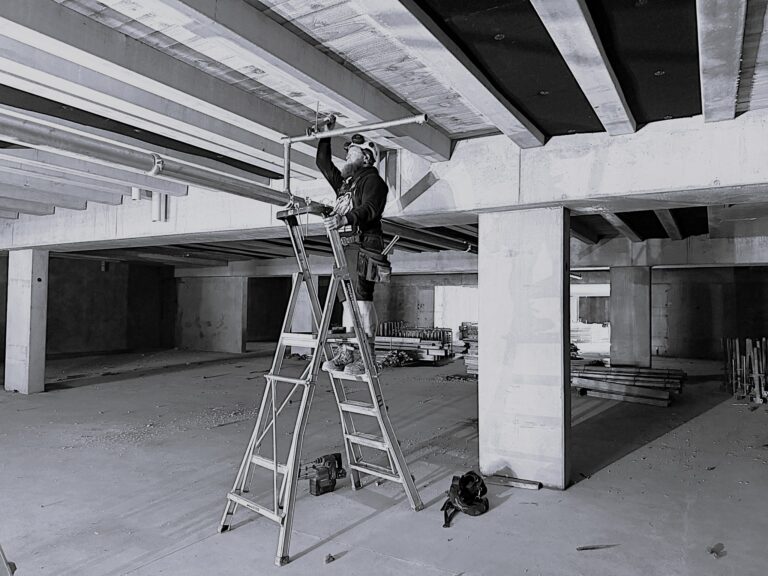 Worker installing a pipe while standing on a ladder.